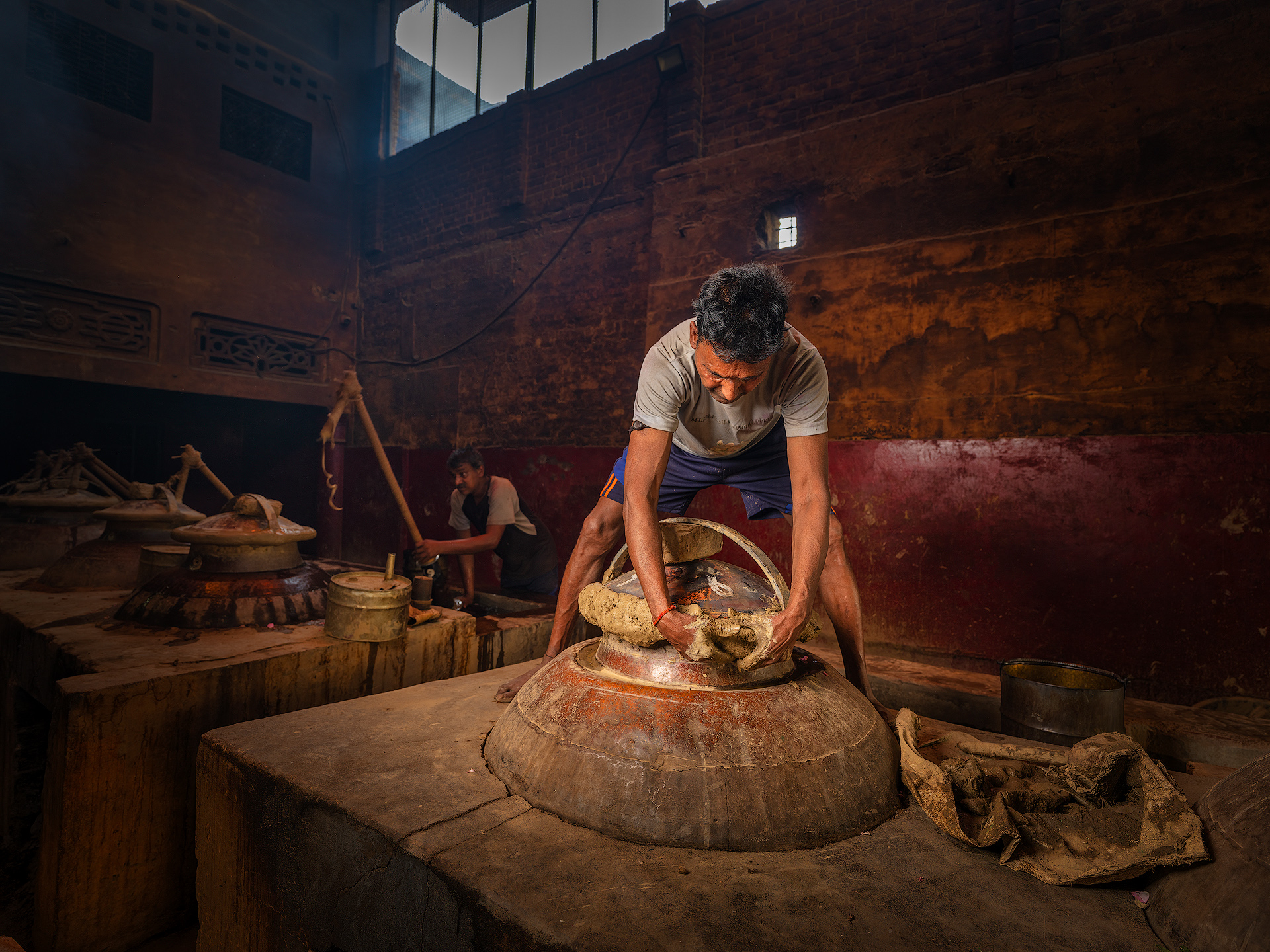 To seal the deg—the copper cauldron used in traditional distillation—a hand-prepared mixture of clay is applied just before the process begins. The clay typically comes from local soil and has a key natural property: plasticity. This means it can be easily shaped and molded without breaking when mixed with water—similar to soft modeling clay or pottery material. The mixture is then applied around the rim of the deg’s lid, along with wet cloth strips, to create an airtight seal. The goal is to prevent the fragrant steam from escaping during distillation, forcing it instead to travel completely through the chonga and into the bhapka, where it condenses. Once the clay dries slightly and hardens with the heat, it forms a very effective natural seal. At the end of the process, a hammer (hathoda) is used to break the now-dry clay crust and open the deg. Distillery Pranav.