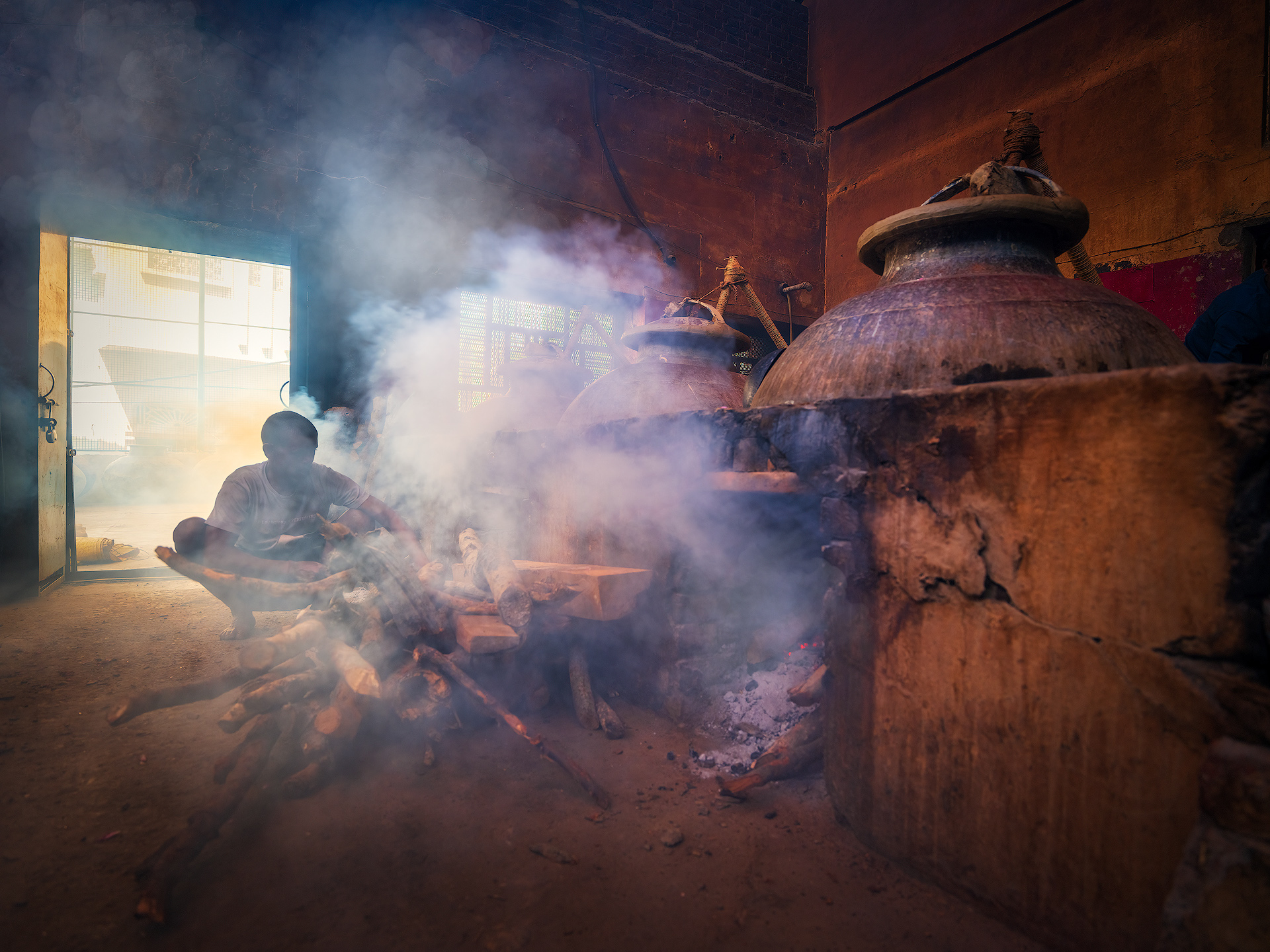 In the traditional attar distillation process in Kannauj, fire control—locally known as bhatti—is a crucial phase often entrusted to an experienced worker called a dighaa. This role requires considerable expertise, as both the temperature and speed of distillation must be manually regulated to ensure the quality of the essence produced.
The bhatti is a traditional furnace built from bricks and clay, typically fueled with firewood or dried cow dung. Its function is to heat the deg, the large copper cauldron where flower petals and water are placed. The heat causes the essential oils to evaporate and travel through the chonga (a bamboo or metal pipe) into the bhapka, where the vapors condense.
The dighaa is responsible for maintaining a steady and appropriate temperature throughout the entire distillation process, which can last several hours. This includes:
• Regulating the heat: Adding or removing fuel to control the intensity of the fire.
• Monitoring condensation: Ensuring the vapor condenses correctly inside the bhapka, preventing overheating or excessive cooling.
• Timely interventions: Using wet cloths to quickly cool the deg if needed, temporarily halting the distillation.
This job demands constant attention and a deep understanding of the process, as even a small variation in temperature can negatively affect the quality of the attar being produced. Distillery Pranav.