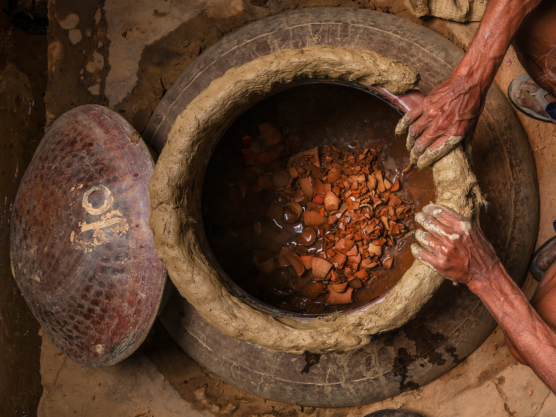 Pranav Distillery. The deg is the central vessel in the traditional attar distillation process in Kannauj, India’s historic “perfume capital.” This large copper cauldron is filled with fresh flower petals—typically roses or jasmine—and water. To prevent any loss of aromatic vapor, the deg is tightly sealed using a mixture of natural clay and wet cloth strips, which is carefully applied around the rim of the lid. This method, handed down through generations, serves to lock in the steam and maintain stable pressure throughout the distillation. As the bhatti (a traditional earthen furnace) heats the deg, the clay seal hardens quickly, forming a strong, airtight bond. The aromatic vapors travel through a connecting pipe (chonga) into the bhapka—a brass or copper receiver submerged in cold water—where the vapor condenses into liquid attar. In the case shown in the photo, the cauldron contains mitti—a special kind of earthy clay sourced from deep layers of soil. The clay is first dried and then baked into small disc-like pieces, similar to tiles. These are used to distill a unique attar known as “mitti attar”, which captures the scent of earth after the first rain—a deeply nostalgic and cherished aroma in Indian culture. Once the distillation is finished, the hardened clay seal is broken with a metal hammer known as a hathoda. This final step allows access to the used petals and reveals the strength and quality of the aromatic essence collected.