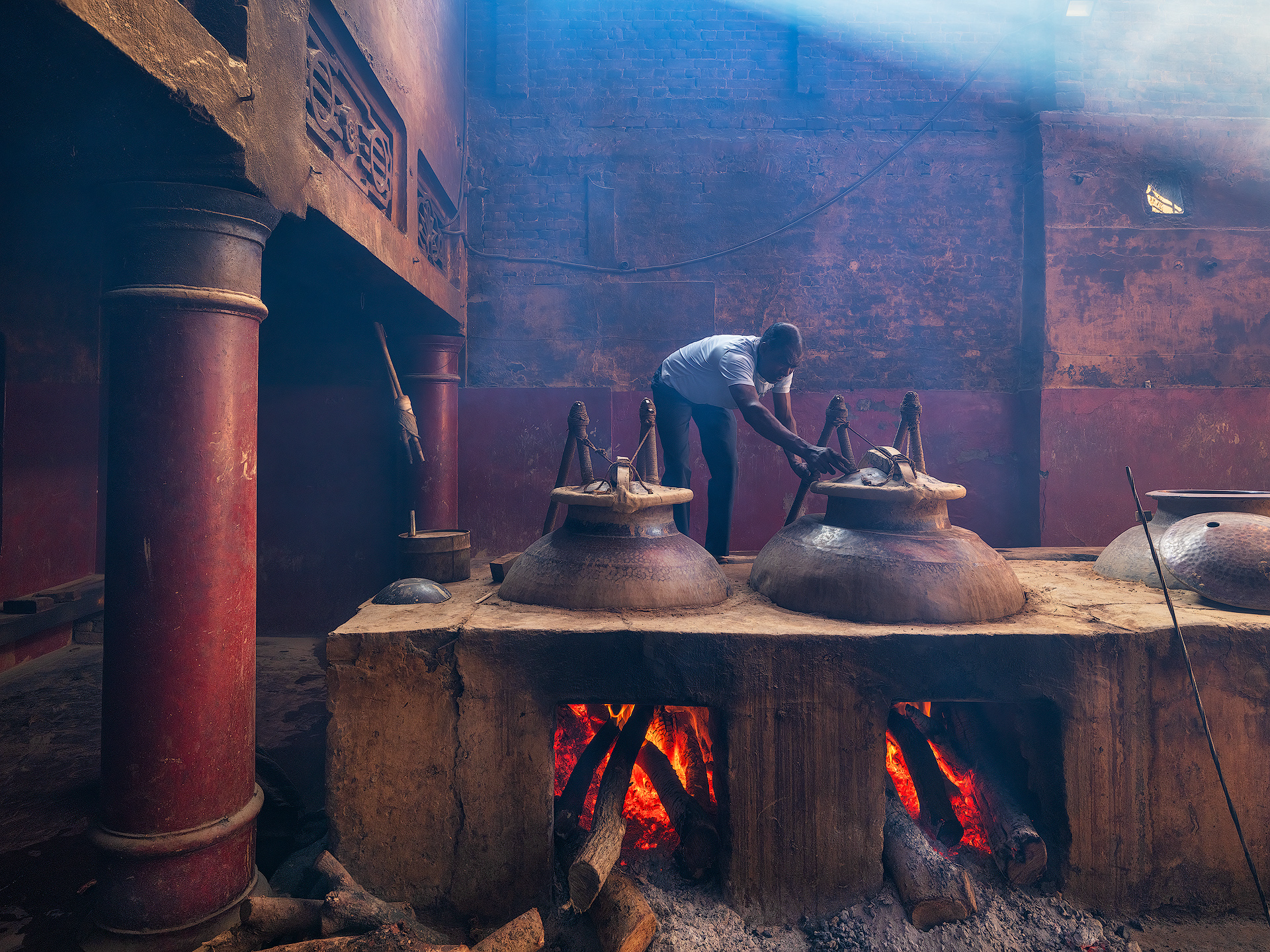 Once the deg is sealed, the distillation process begins. A worker remains on watch to monitor each phase of the procedure.
Deg (or Degah): A large copper cauldron where rose or jasmine petals are mixed with water for distillation.
Fire (or Bhatti): The heat is provided by a traditional earthen furnace called a bhatti, fueled by wood or dried cow dung, which heats the deg causing the water and aromatic oils to evaporate.
Bhapka: A metal receiver, usually made of brass, where the fragrant vapors condense into liquid. It is partially submerged in cold water to cool the vapor.
Chonga: A bamboo or metal pipe that connects the deg to the bhapka, allowing the aromatic vapor to travel during distillation.
