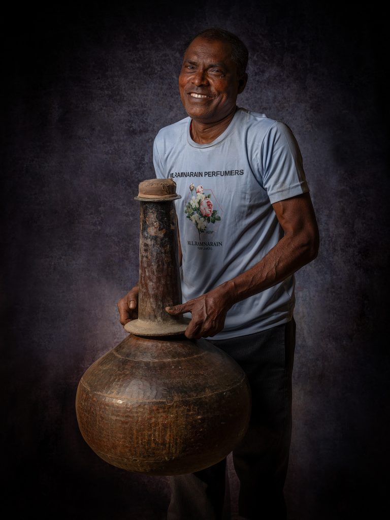 A worker with the bhapka:
A metal receiver, usually made of brass, where the fragrant vapors condense into liquid. It is partially submerged in cold water to cool the vapor during the traditional distillation process.